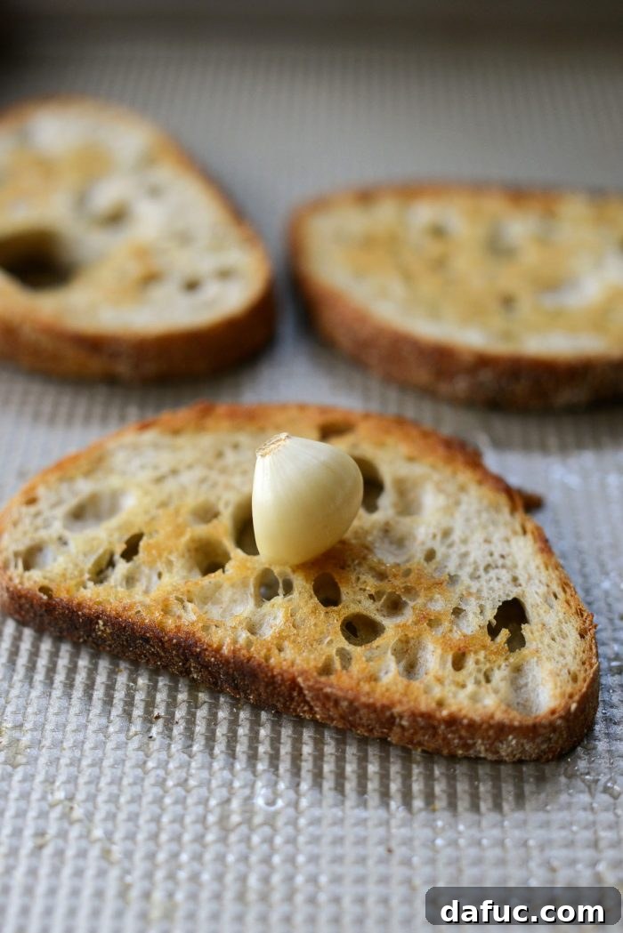 Hands demonstrating how to lightly rub a peeled garlic clove over a slice of warm, toasted bread, imparting subtle garlic flavor.