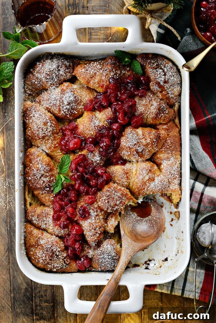 A slice of Eggnog Croissant French Toast Bake topped with powdered sugar, cranberry relish, and maple syrup on a plate, with the full casserole in the background.