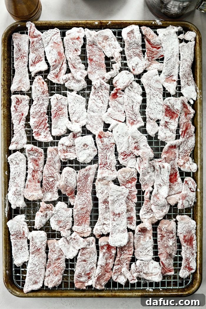 Cornstarch-coated beef strips meticulously arranged on a wire rack, which is set over a rimmed baking sheet.