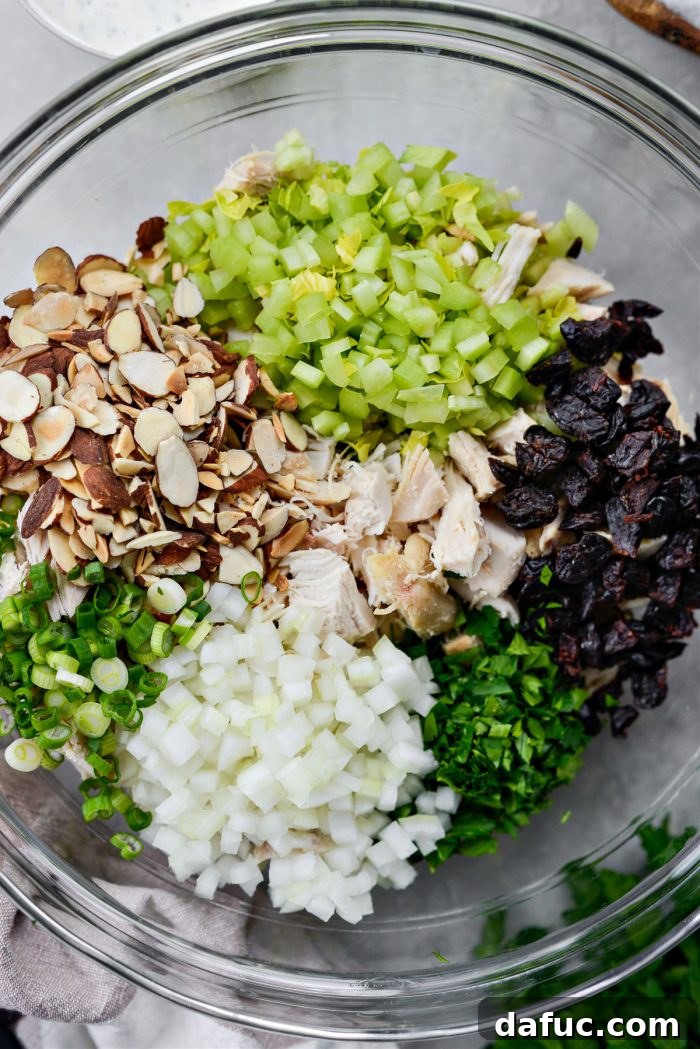 All prepped salad ingredients including shredded chicken, celery, onions, dried cherries, and toasted almonds in a large mixing bowl.