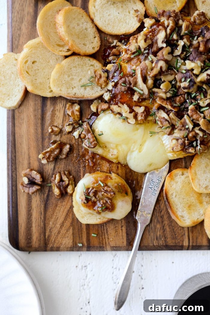Full shot of the Honey Baked Brie with fig jam, walnuts, rosemary, and crostini on a wooden board.