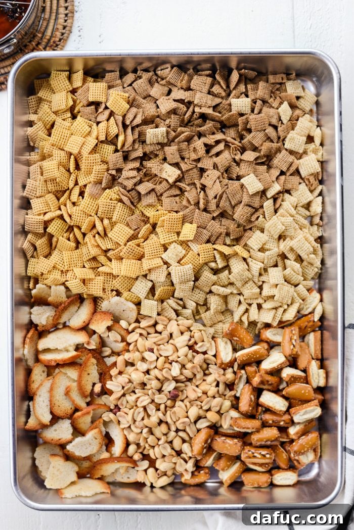 The Ultimate Everything Bagel Chex Mix 6 Variety of dry ingredients for Chex Mix in a large deep-sided pan: corn, rice, and wheat Chex cereals, broken plain bagel chips, sourdough pretzel nuggets, and roasted peanuts.