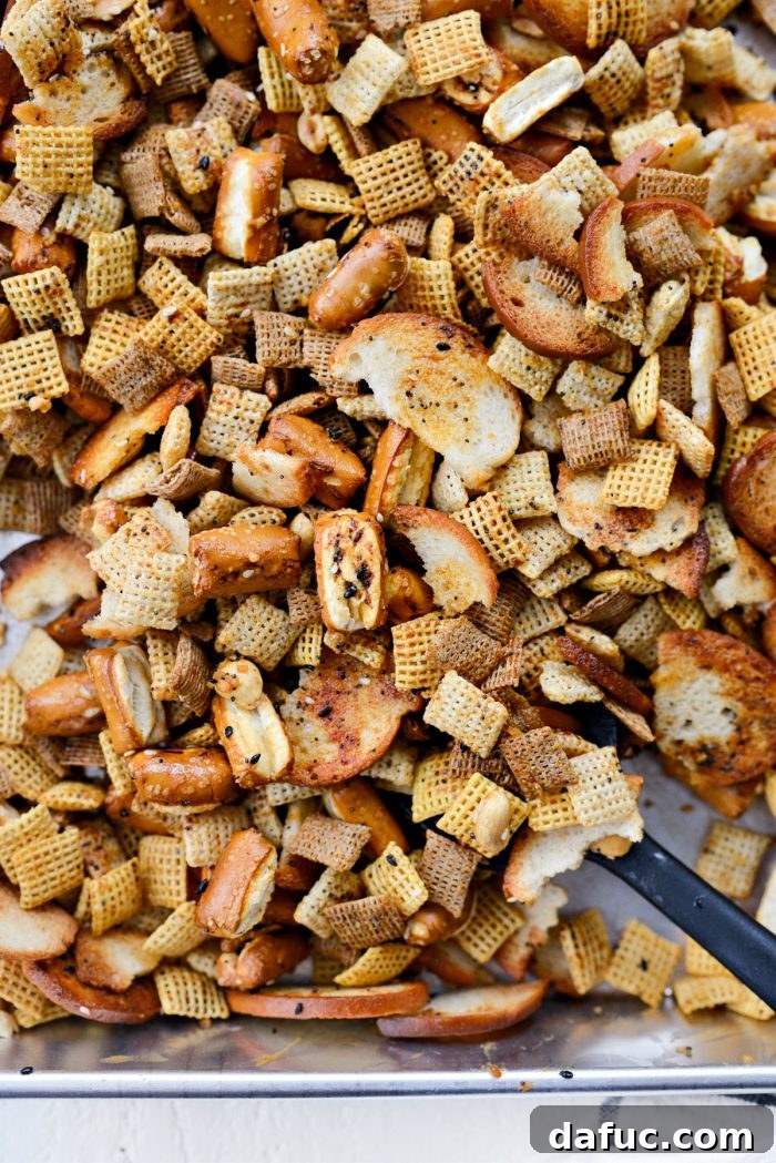 The Ultimate Everything Bagel Chex Mix 9 A close-up of the Chex Mix being tossed vigorously with a spatula in a deep pan, ensuring the butter and seasoning mixture thoroughly coats all the cereal, bagel chips, pretzels, and peanuts.