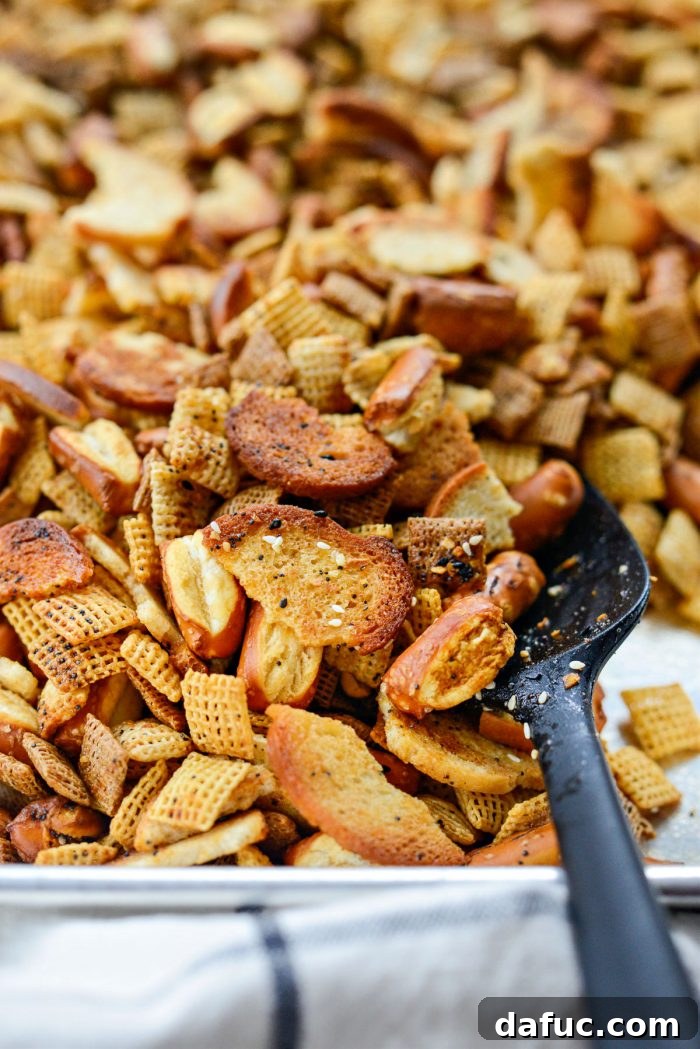 The Ultimate Everything Bagel Chex Mix 10 Everything Plus the Bagel Chex Mix baking in the oven, spread out on a sheet pan, with a spatula ready for stirring every 15 minutes to achieve an even golden crispness.