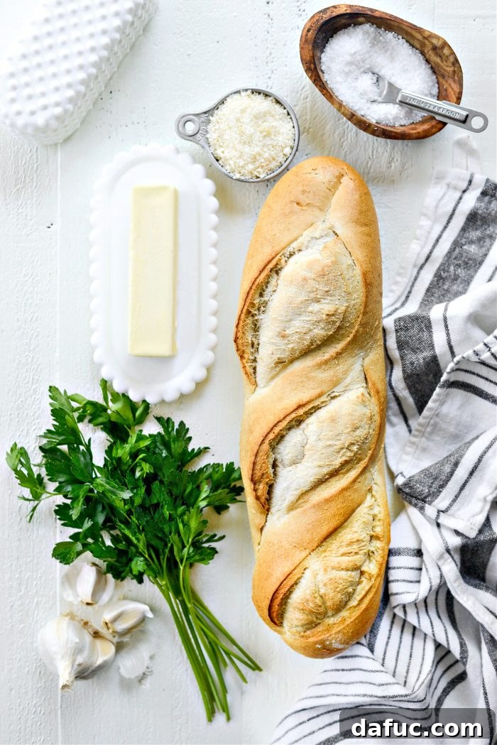 A selection of fresh ingredients laid out for 15-Minute Garlic Bread: a loaf of Italian bread, garlic cloves, softened butter, kosher salt, parmesan cheese, and fresh parsley