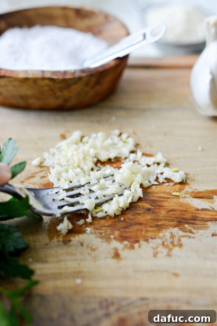 A fork mashing minced garlic and kosher salt on a cutting board, creating a smooth garlic paste