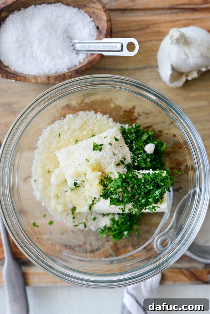 A mixing bowl containing softened butter, garlic paste, parmesan cheese, and fresh parsley, ready to be combined